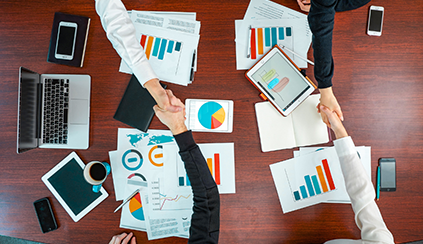 Photo of four people shaking hands over a table covered in charts, graphs, and devices.