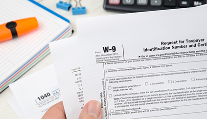 Photo of W-9 tax paperwork held above a desk with a notebook, binder clips, and a calculator