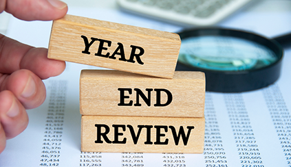 A hand with a stack of blocks that say, "Year End Review" on top of paper with number lists and a magnifying glass and calculator